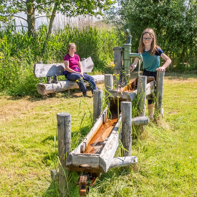 Agnes probeert een waterpomp uit, Ellen zit op een bankje.