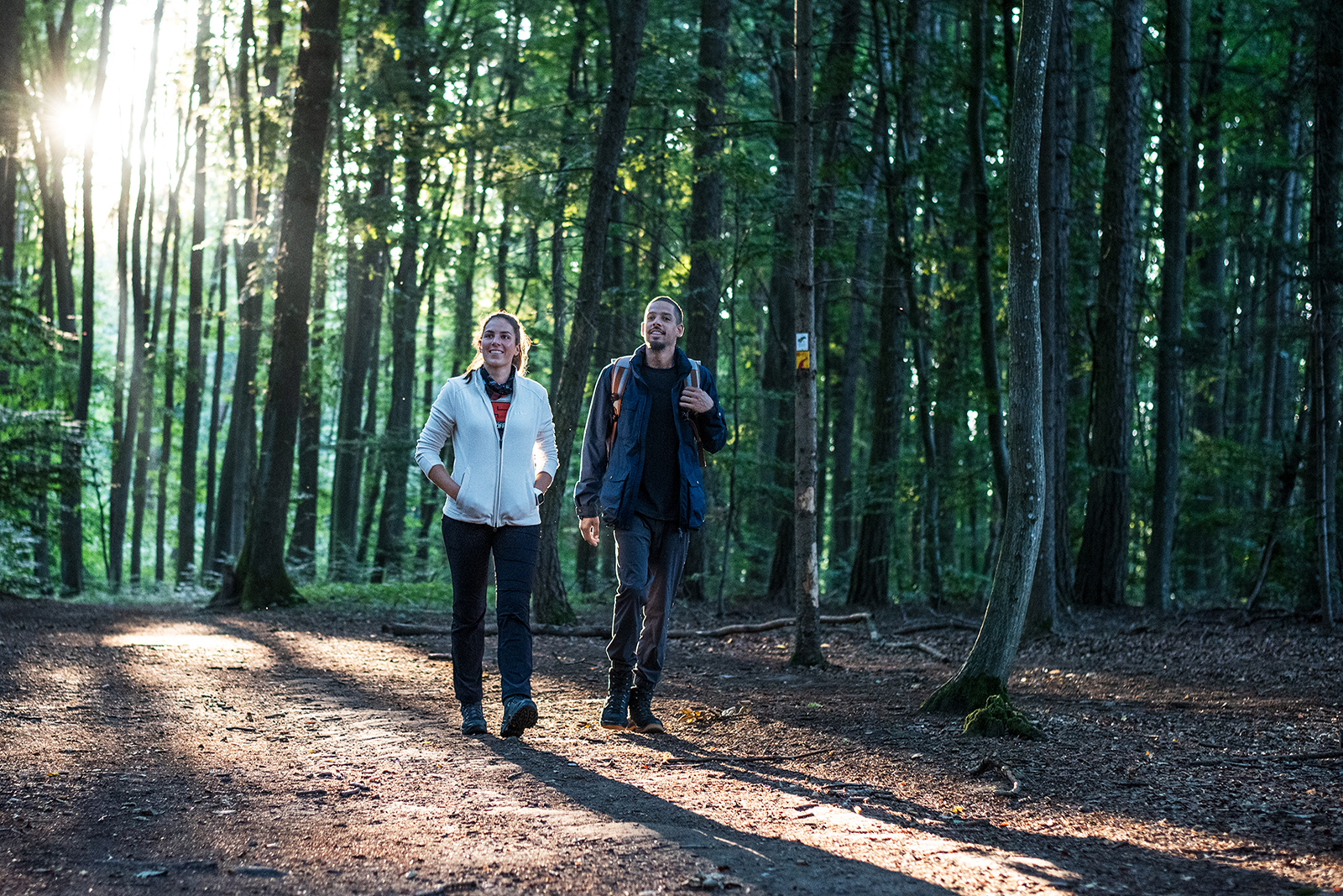 man en vrouw wandelen in het bos met de zon die door de bomen komt.