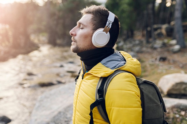 man met gele jas en koptelefoon op die buiten met zijn ogen dicht geniet van de muziek.