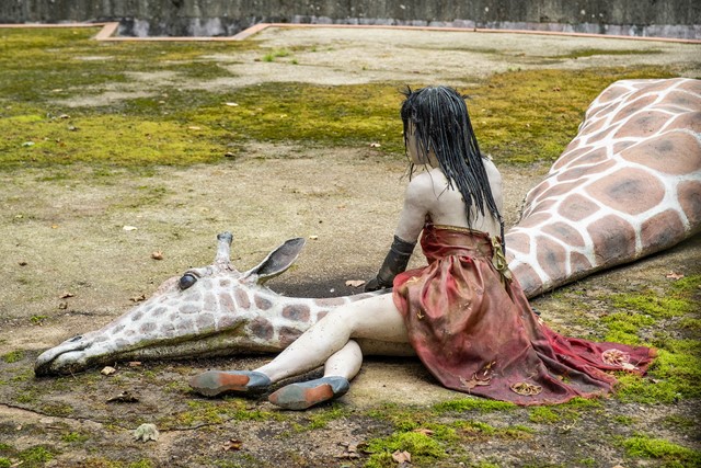 Artistiek beeld in de Berenkuil in Maastricht van een figuur in een rood gewaad die naast de nek van een liggende giraffe zit.