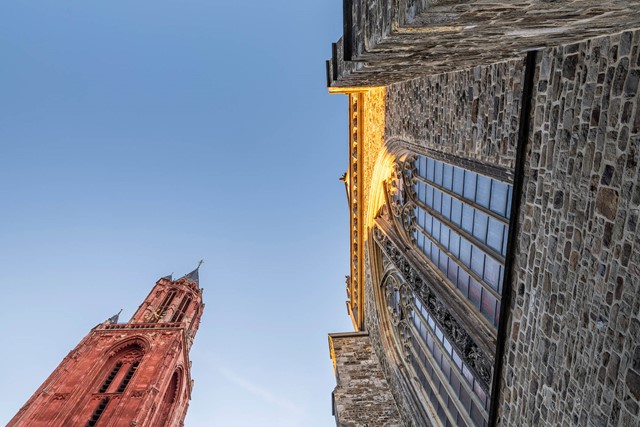 Kikkerperspectief van de Sint-Janskerk in Maastricht, waarbij de kerktoren en een zijkant van het gebouw met grote glas-in-loodramen tegen een helderblauwe lucht worden getoond