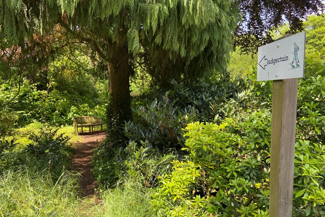 bordje dat wijst naar de Ludgertuin met groene boom en planten en een bankje.