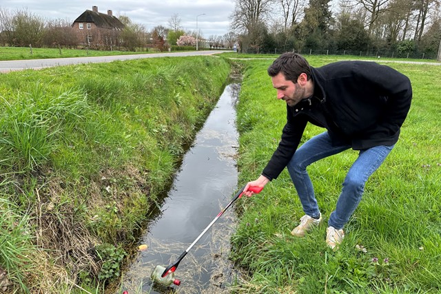 man met afvalgrijpstok grijpt een fles uit een sloot.
