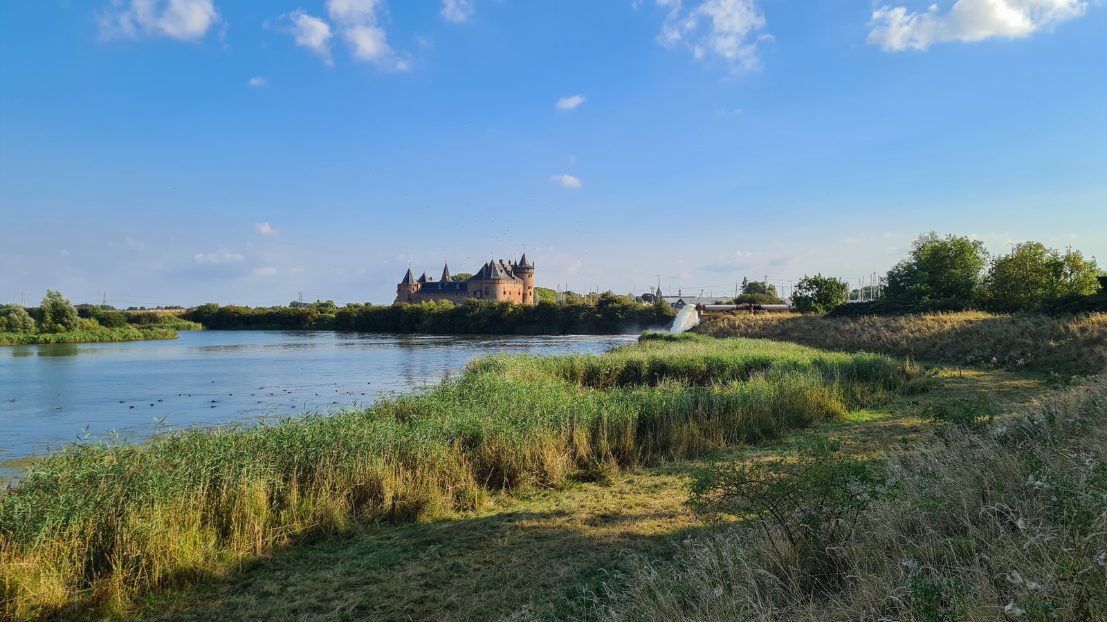 afbeelding van het Muiderslot aan het water en de dijk, met blauwe lucht.
