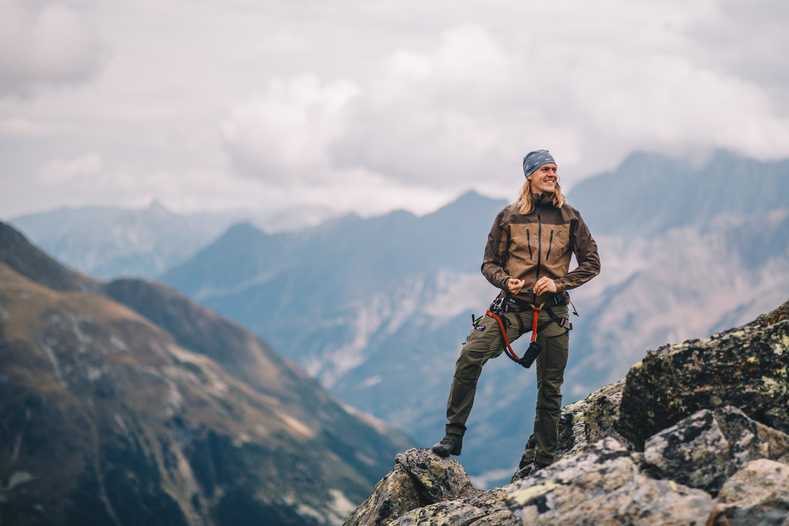 afbeelding van een man met lange haren bovenop een berg met uitzicht op gebergte en wolken.