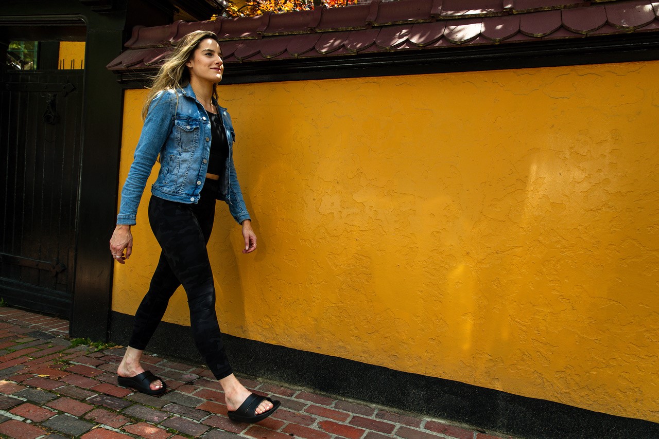 vrouw met OOFOS herstelslippers loopt langs een gele muur.