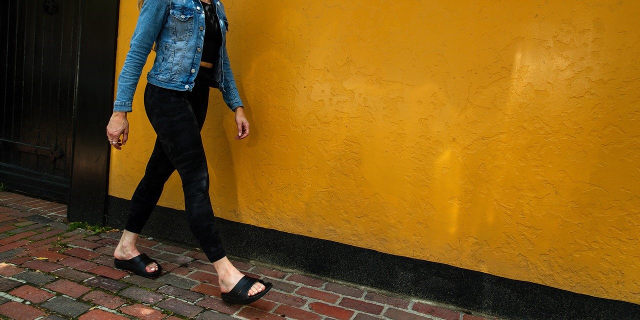 vrouw met OOFOS herstelslippers loopt langs een gele muur.