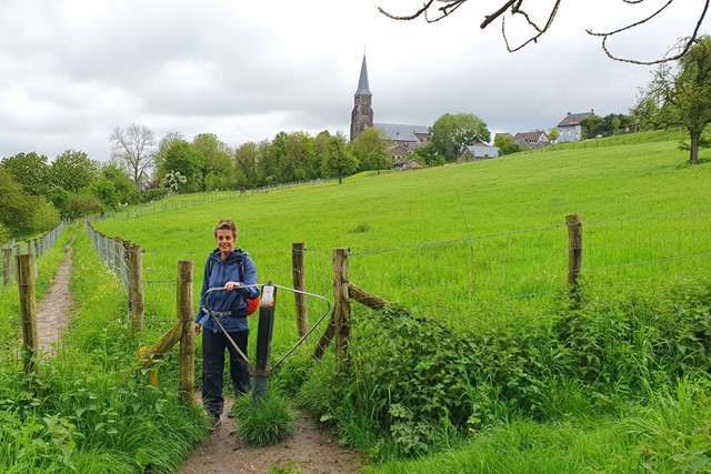 wandelaar op glooiend grasveld met kerk op de achtergrond, bij Vijlen.