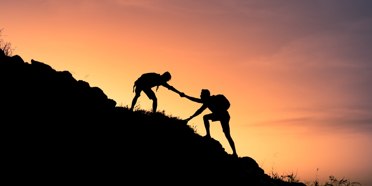 afbeelding van twee wandelaars die elkaar helpen naar de top van een berg.
