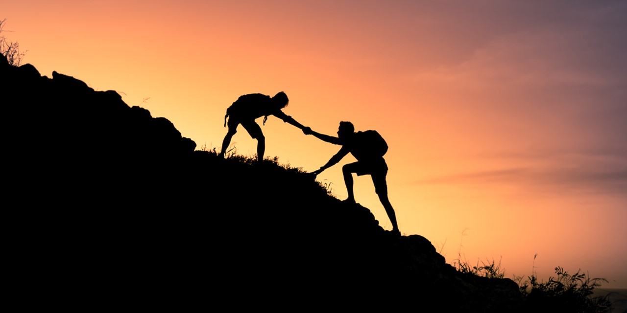 afbeelding van twee wandelaars die elkaar helpen naar de top van een berg.
