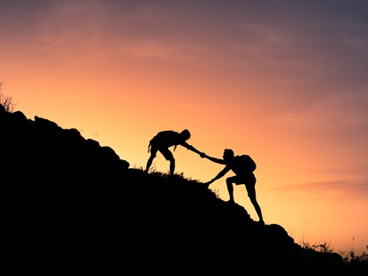 afbeelding van twee wandelaars die elkaar helpen naar de top van een berg.